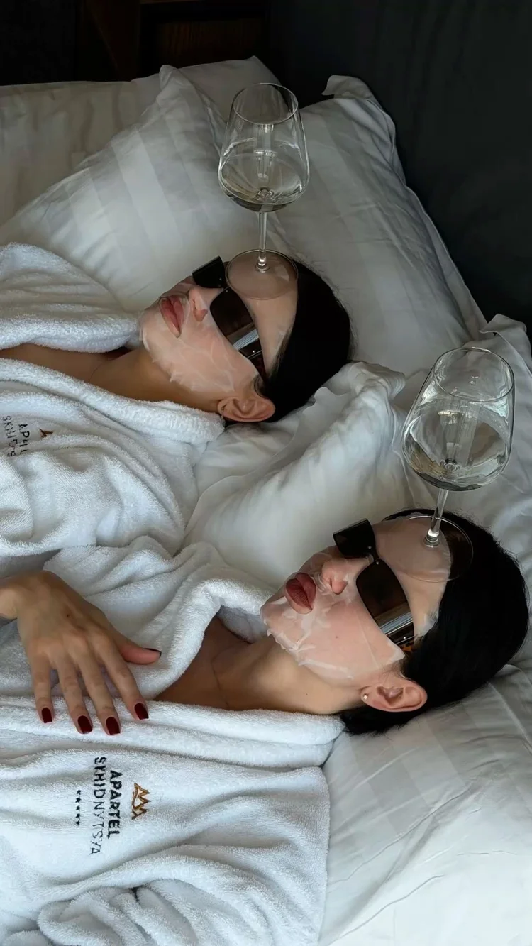 Two of the girls in bathrobes and face masks, balancing wine glasses on their sunglasses