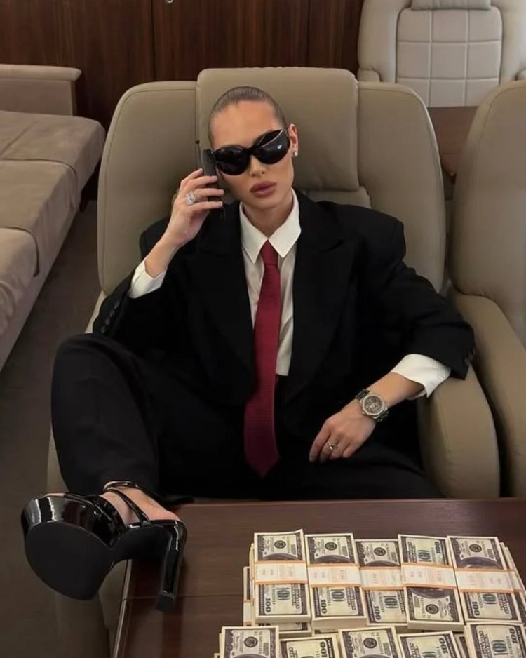 A woman in a black suit and sunglasses on the phone in a private jet, stacks of cash on the table