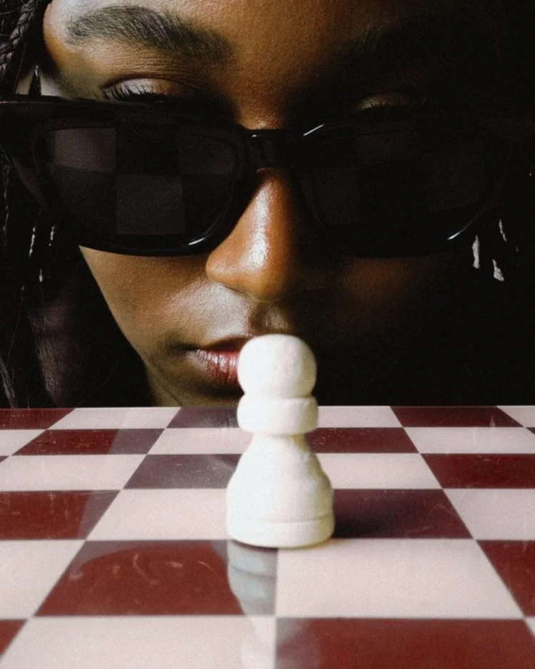 A woman in sunglasses looking over a single white pawn on a burgundy and white chessboard
