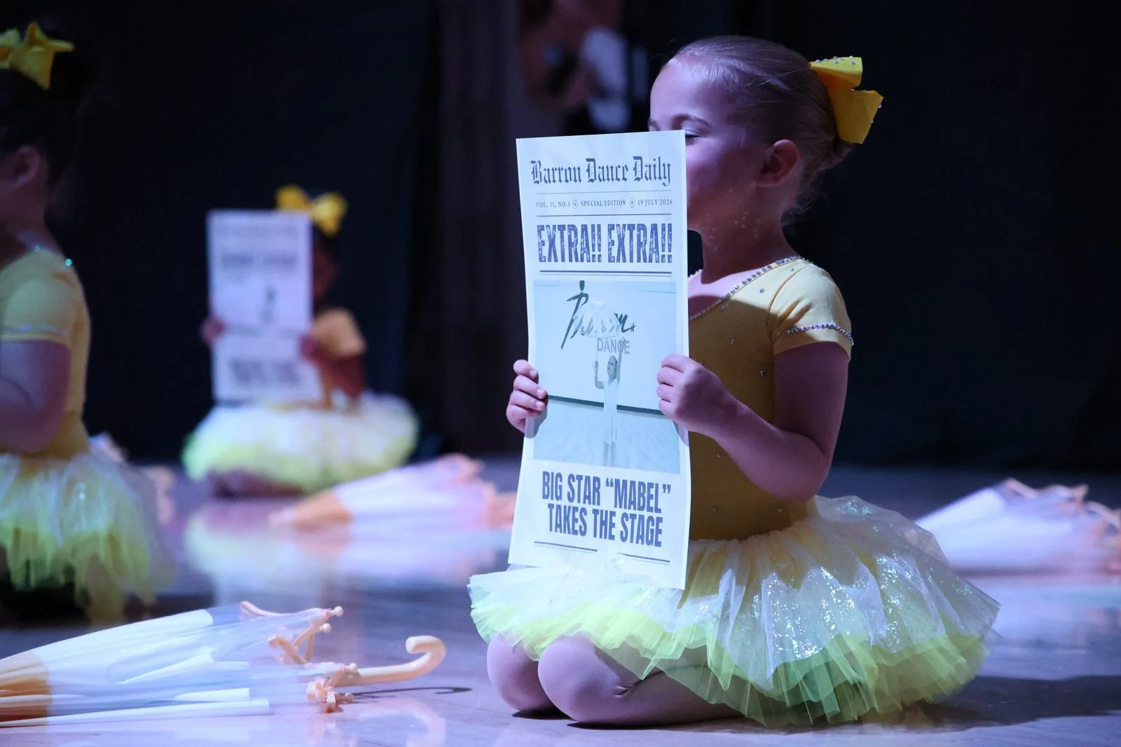 A young dancer on stage at the first Barron Dance recital holding a Barron Dance Daily newspaper prop