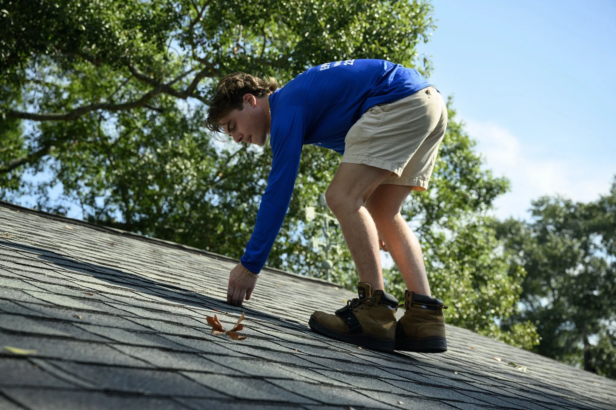 Epic Roofing crew member crouched on a shingle roof slope under tall trees