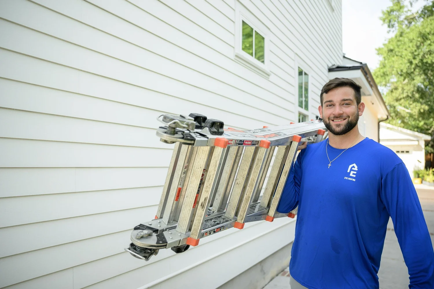 Epic Roofing crew member smiling with an aluminum ladder on his shoulder beside a white house