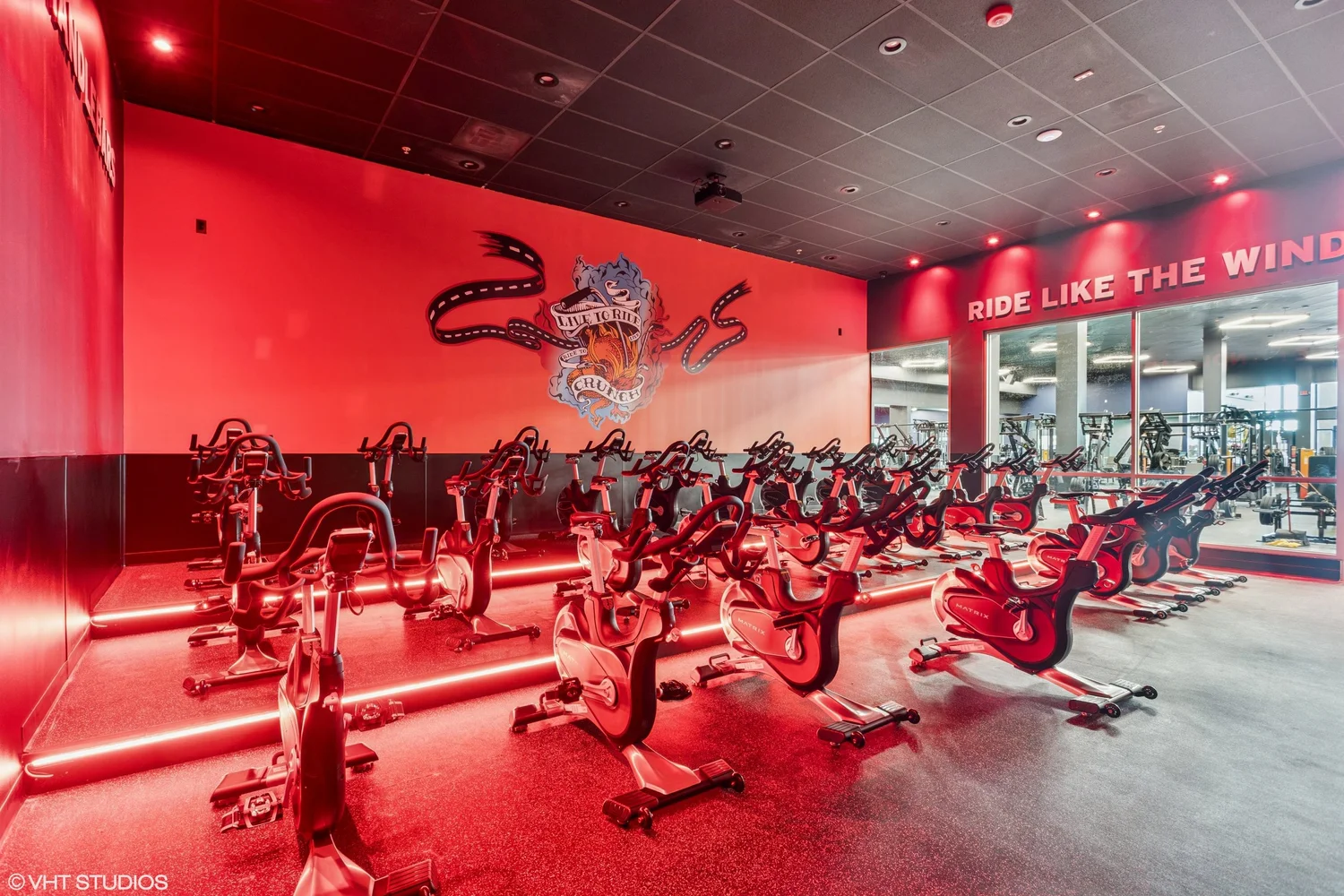 The cycling studio lit entirely in red, rows of bikes facing a mirrored wall under Ride Like The Wind.