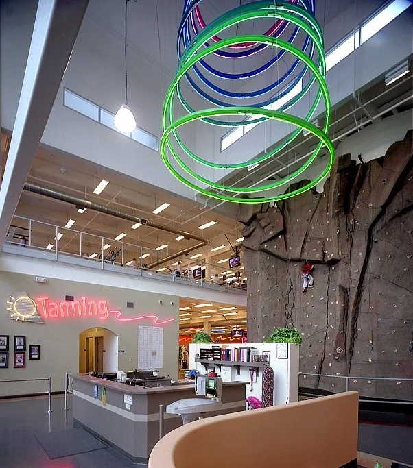 The front desk under the neon rings, a curved pale timber counter with the climbing wall rising behind.