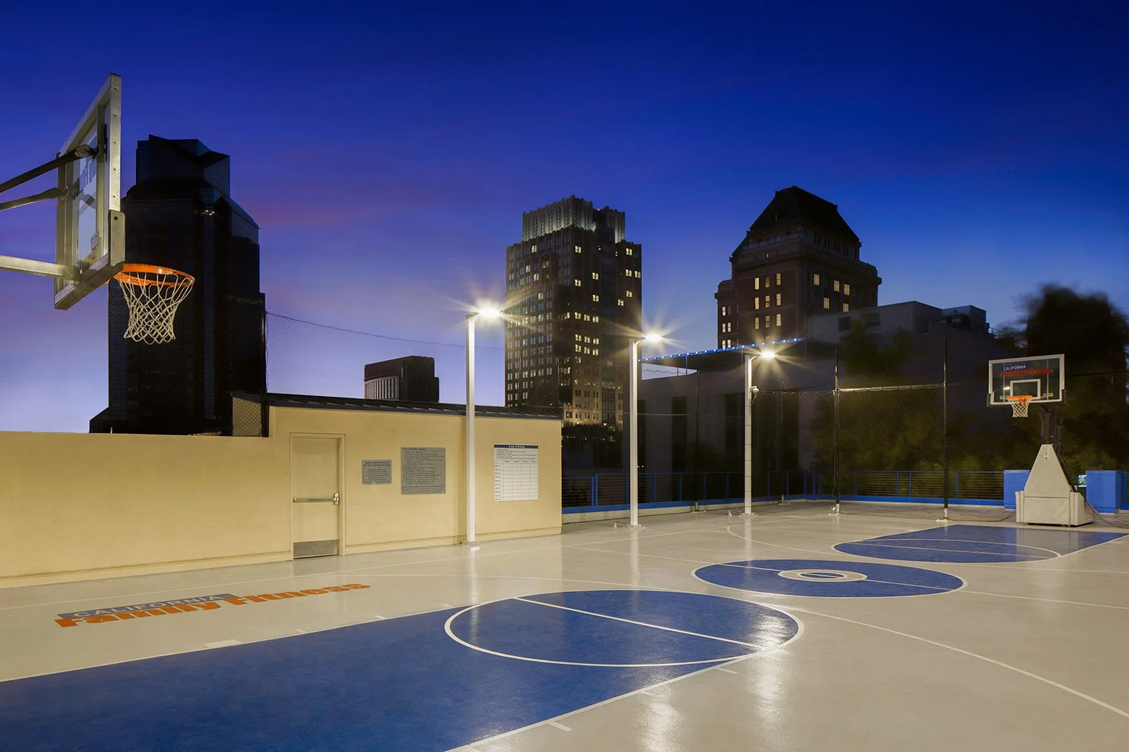 The same court after dark under floodlights, the deck lit pale against a deep blue sky with the towers in silhouette.
