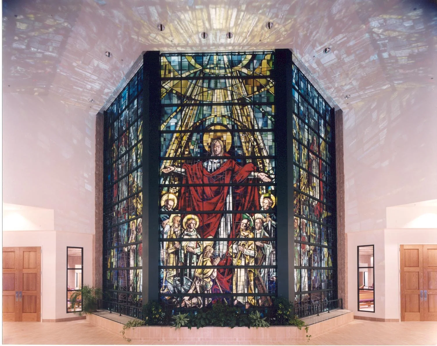 The stained glass wall head on, a robed figure with arms outstretched above haloed saints, between two panelled doors.