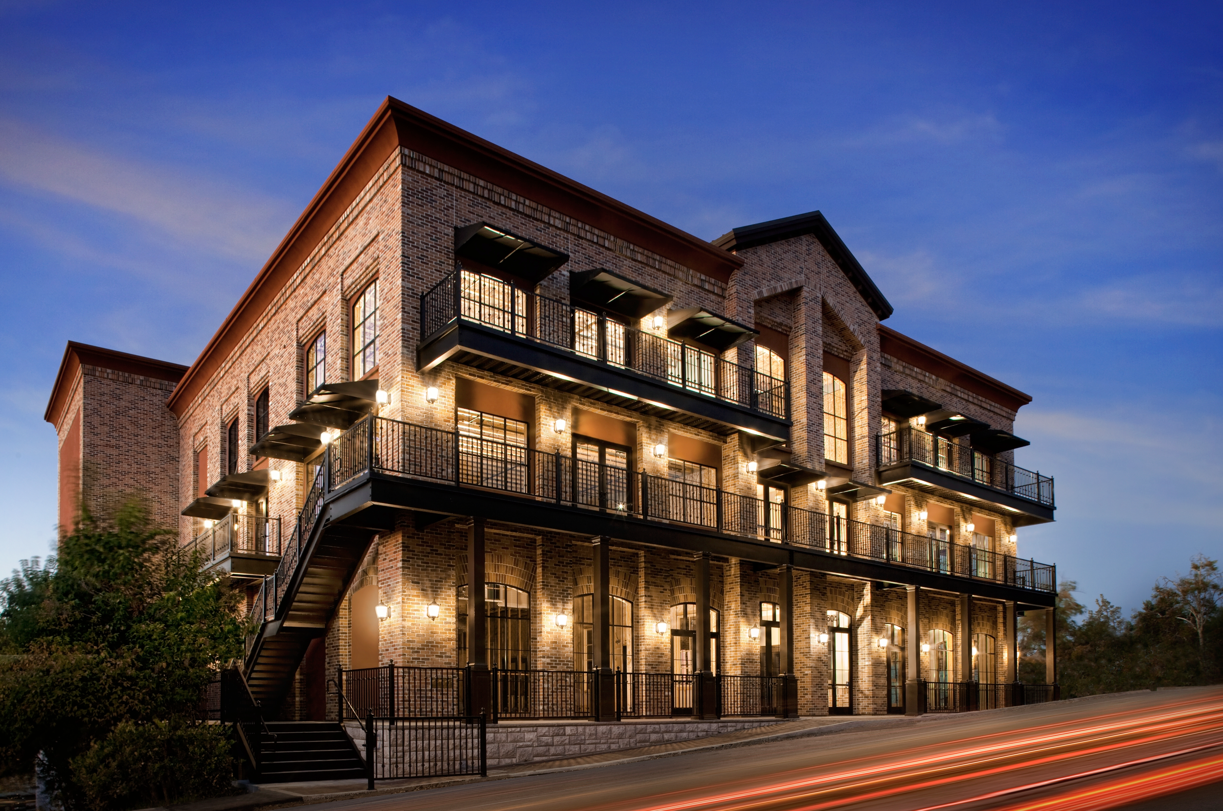 The building on its corner at dusk, three brick storeys on a pale stone base, balconies above lit arched shopfronts.