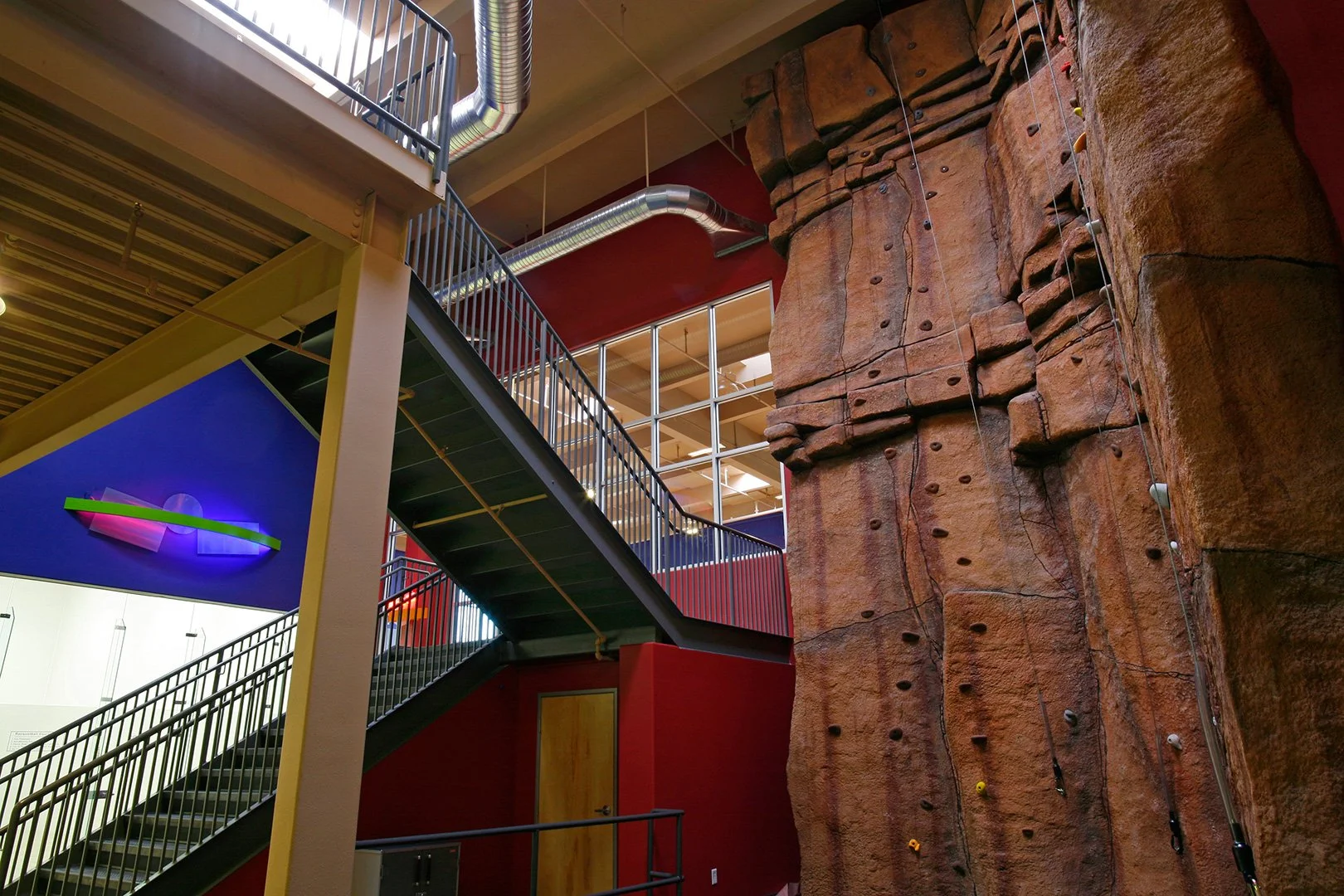 The climbing wall from below, its overhangs rising out of frame behind the open steel stair to the mezzanine.