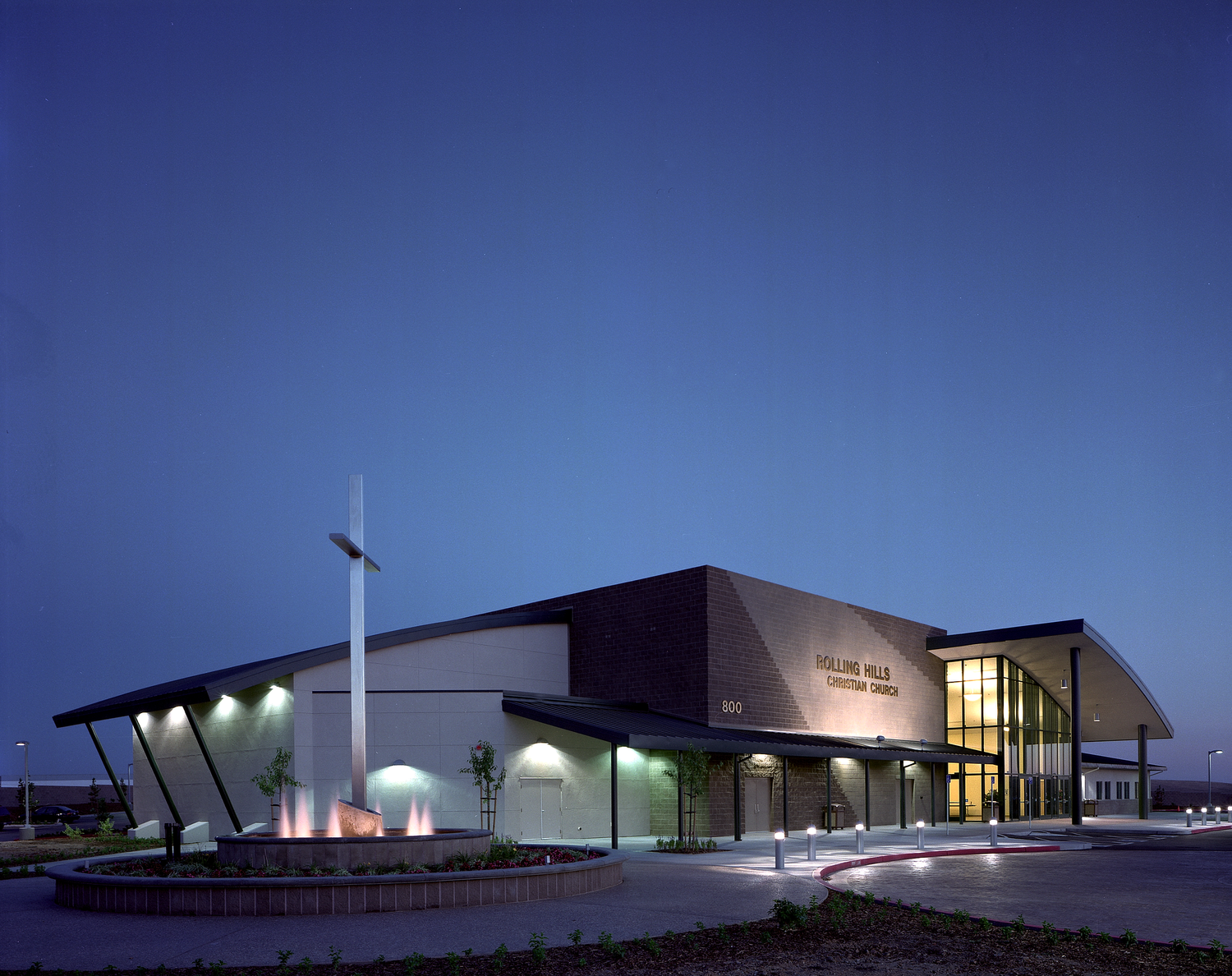 The whole building from the entry drive at dusk, windows lit against a deep blue sky, the cross and fountain at the left.