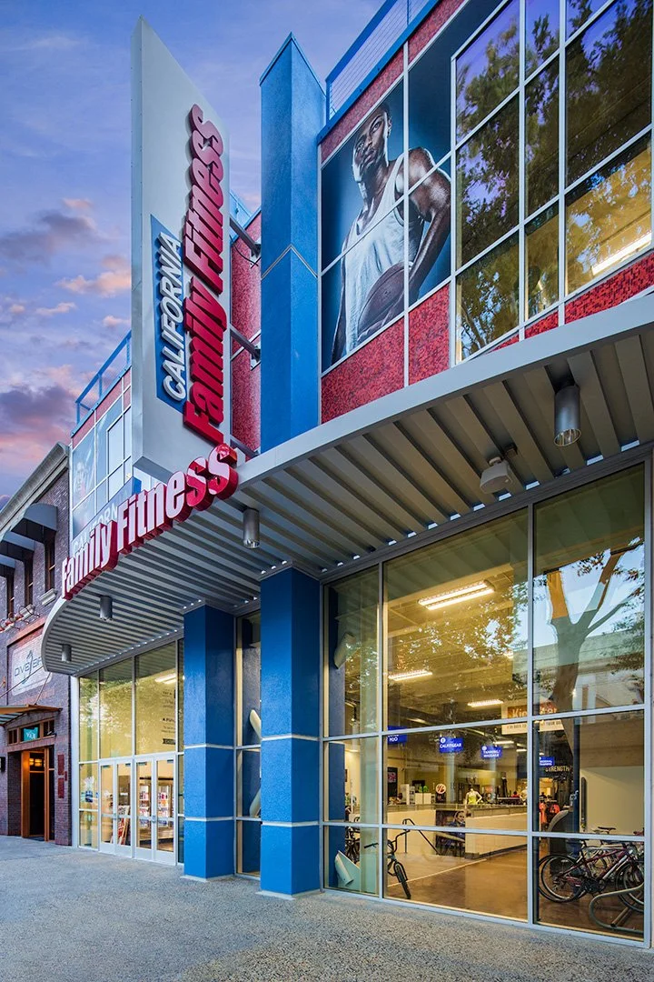 The frontage from further along at dusk, the red lettering on the canopy and blade sign, bikes and a lit gym floor behind the glass.