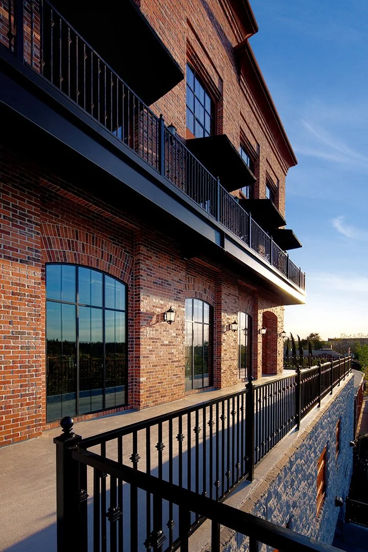 Along an upper balcony into low sun, tall arched windows and lanterns on the left, black railing running away.