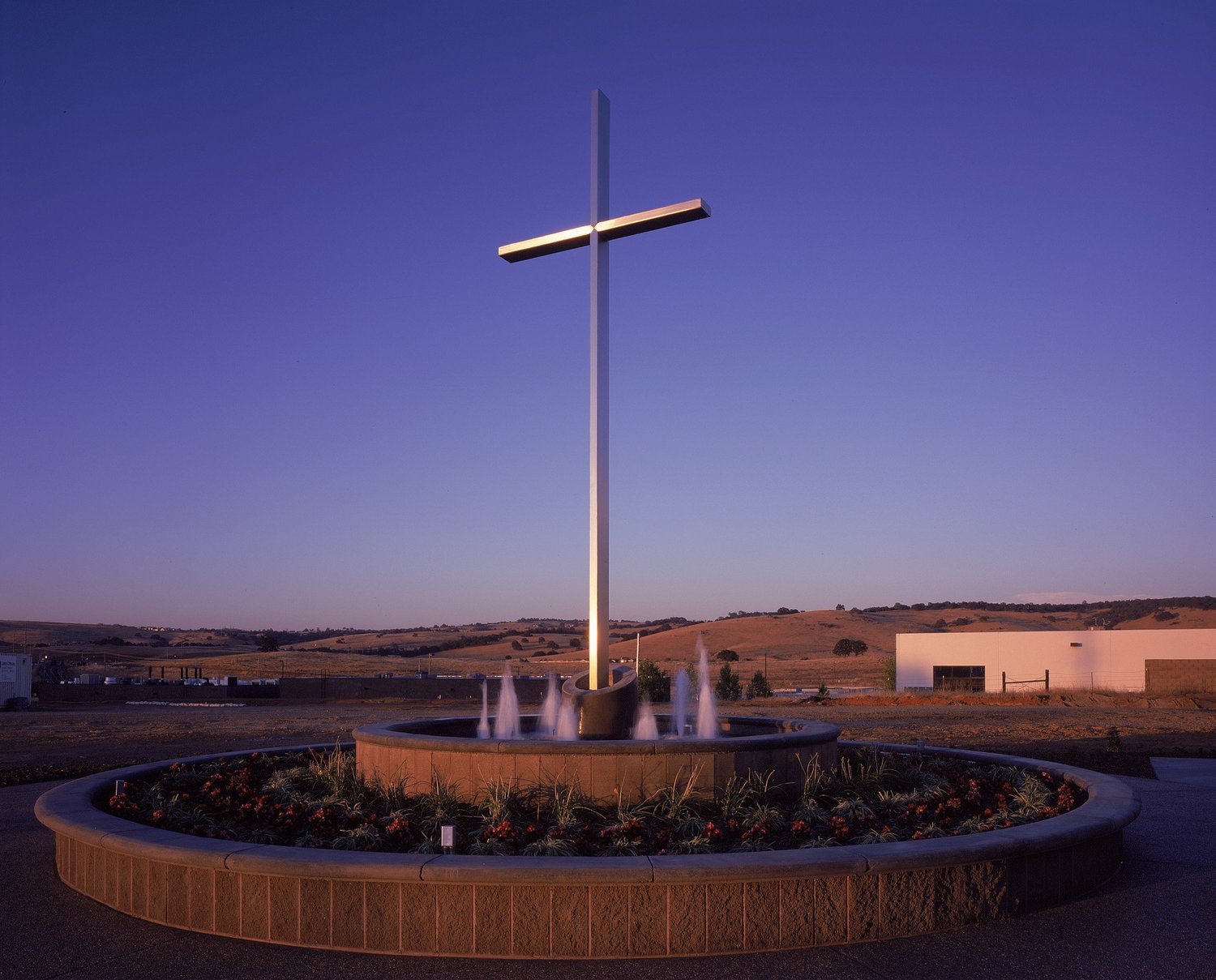 A tall slender cross lit from below over a circular fountain at dusk, open hills beyond.