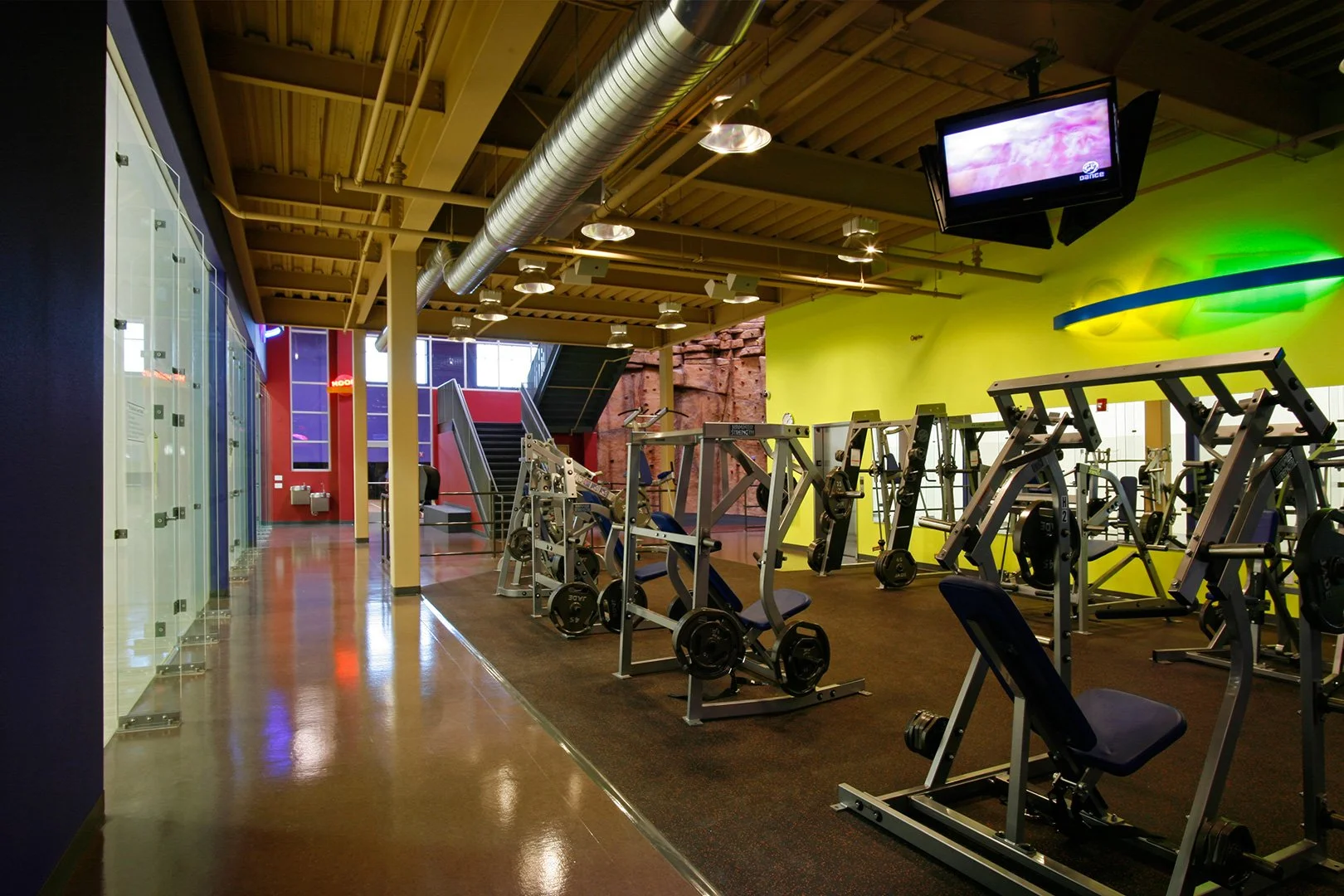Along the free weight floor, plate loaded machines and benches against a yellow wall, the climbing wall beyond.