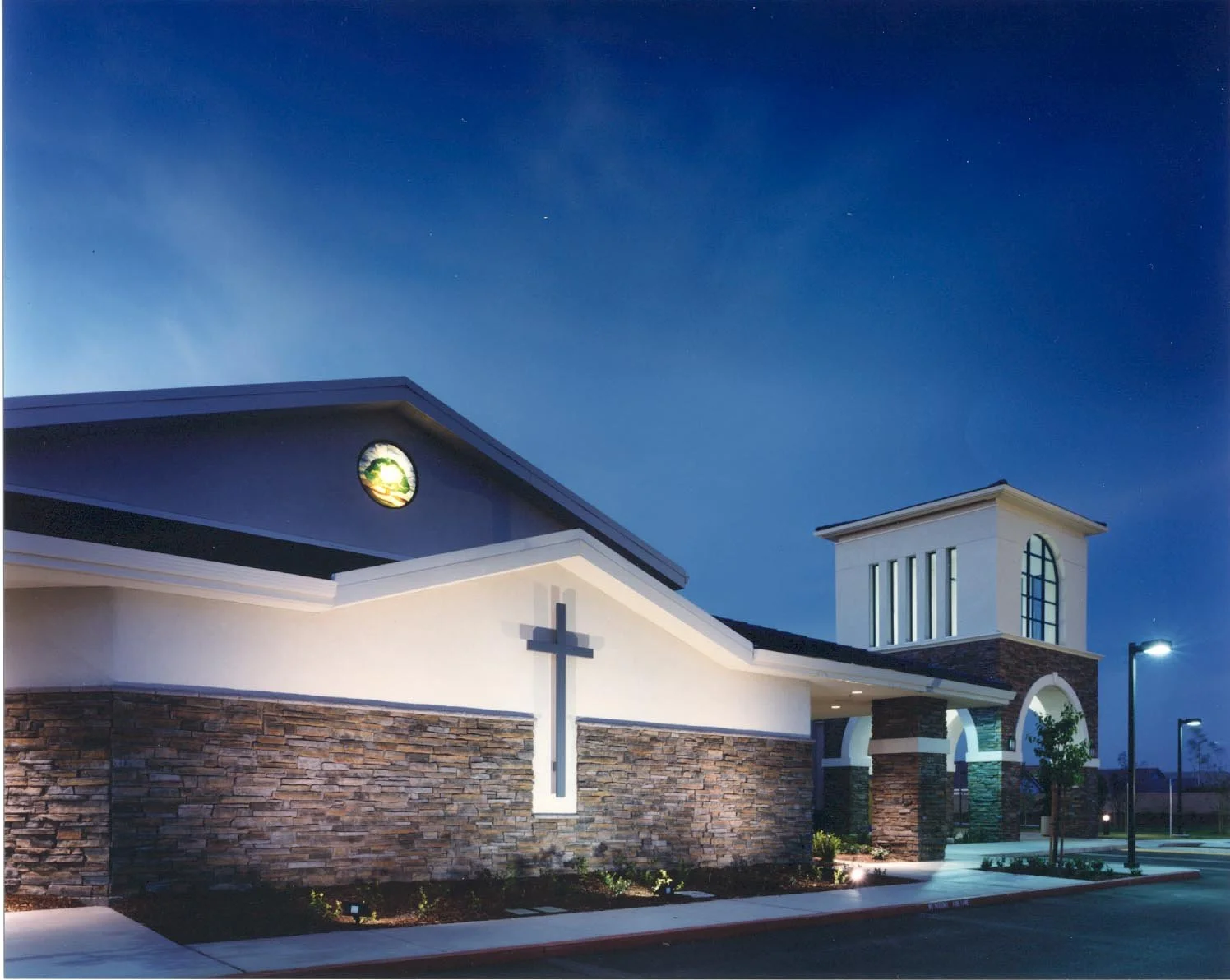 A cross on the white gable above stacked stone, a lit round window beneath, the entry tower beyond.