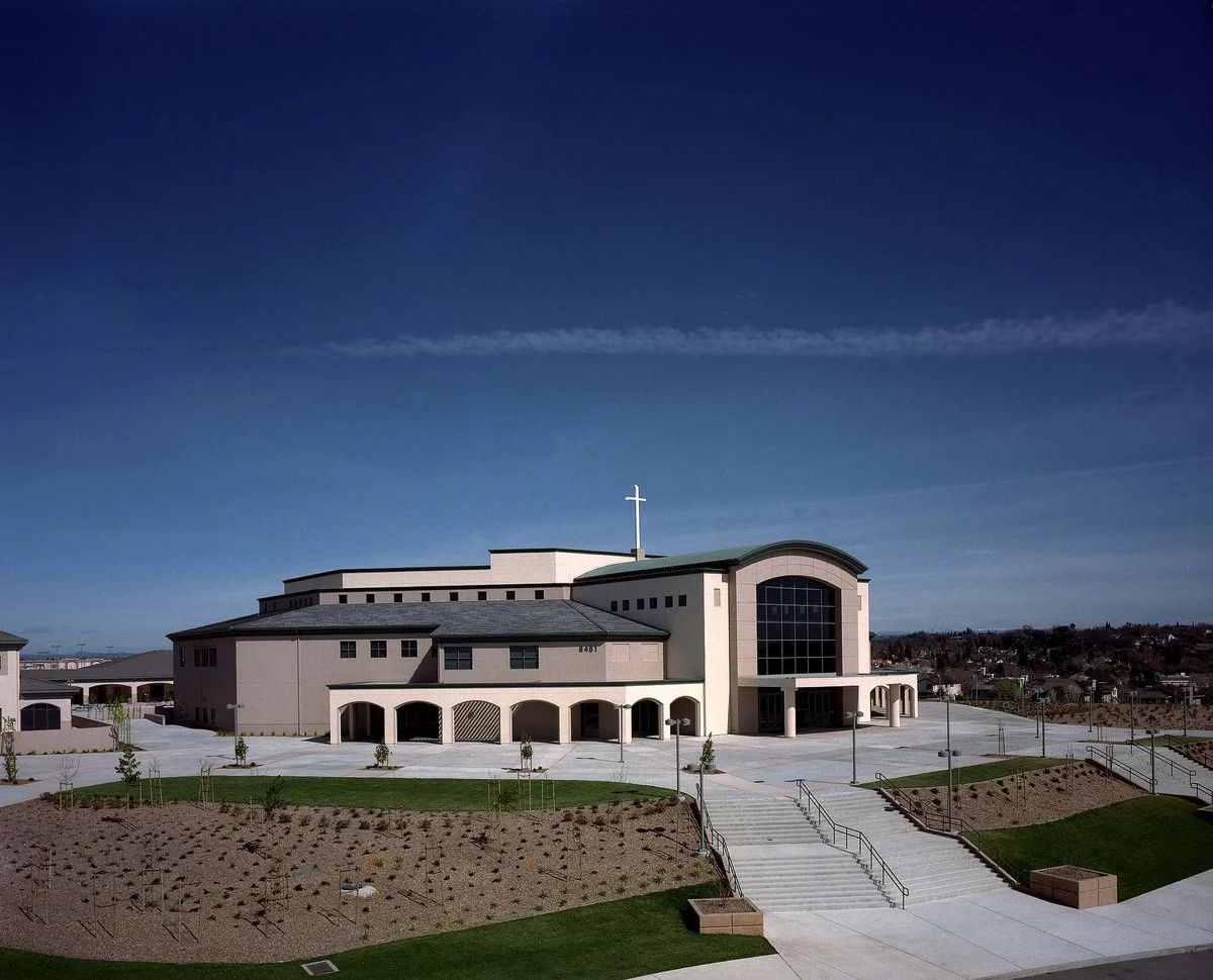 The campus in daylight from across the hill, its arcade running the ground floor below a white cross on the roof.