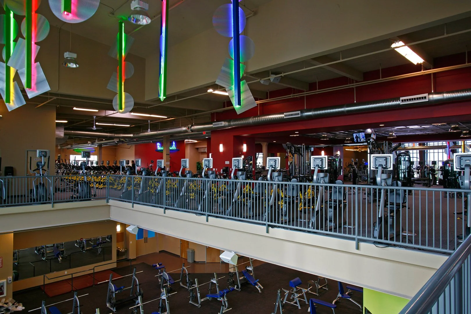 The floor from the mezzanine, a long rank of cardio machines behind a steel rail against a red wall.