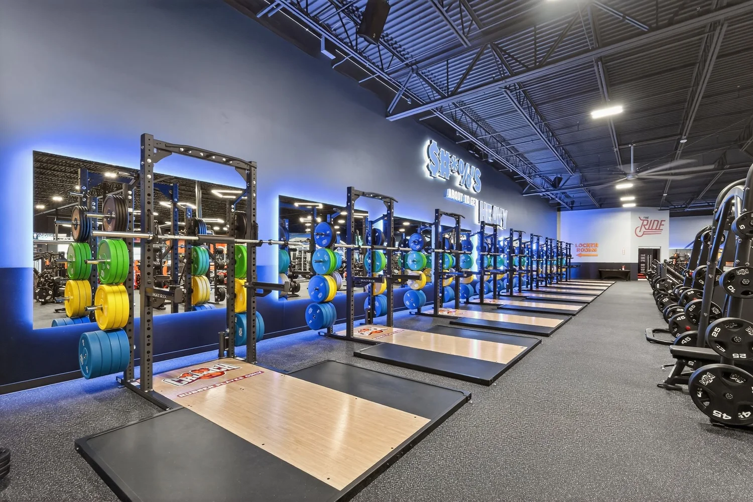 Squat racks and lifting platforms down one side of the club under a blue-lit wall.