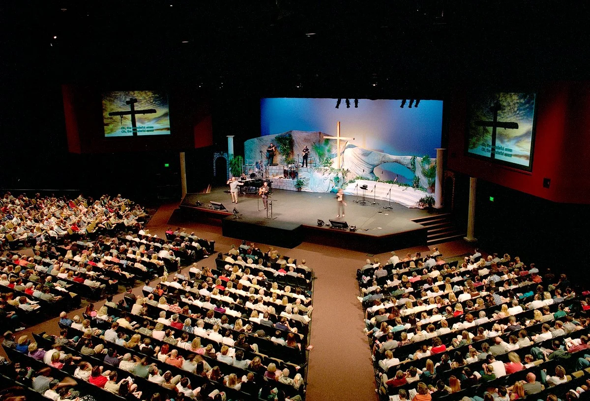 The auditorium during a service, a full house of raked seating facing a band and a tall timber cross.