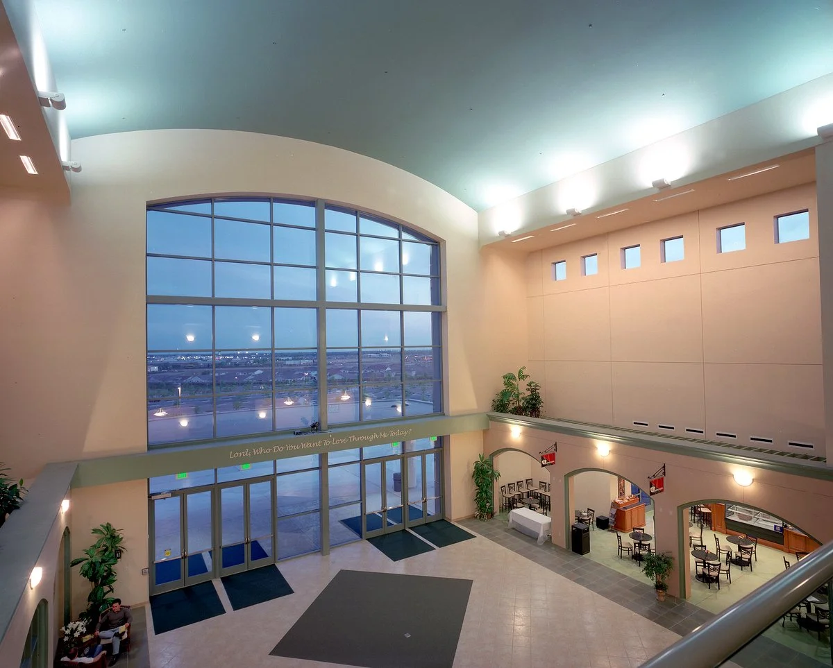 The upper lobby at dusk, a two storey arched window filling the end wall above the cafe tables below.