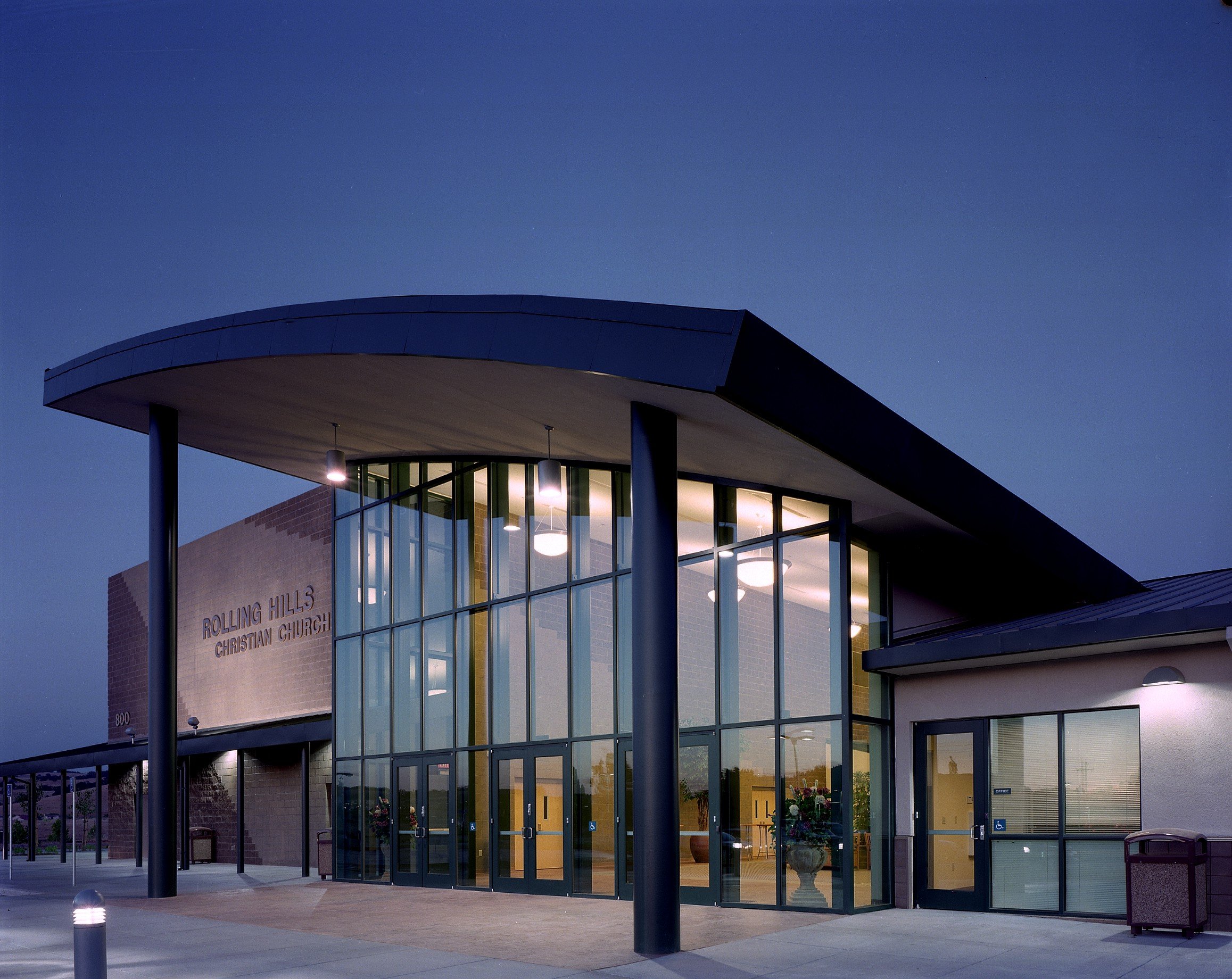 The church entrance at dusk, a long curved canopy on slender dark columns over a lit two storey glass wall.