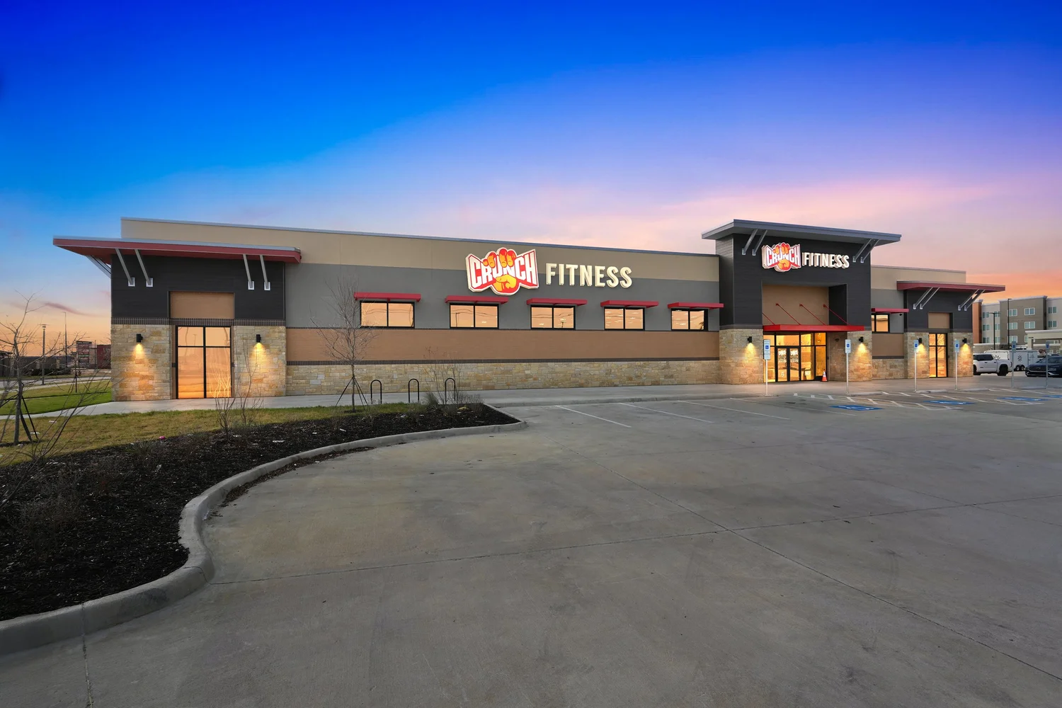 The club at dusk, a long facade of grey panel and stone with red canopies and the lit Crunch Fitness sign.