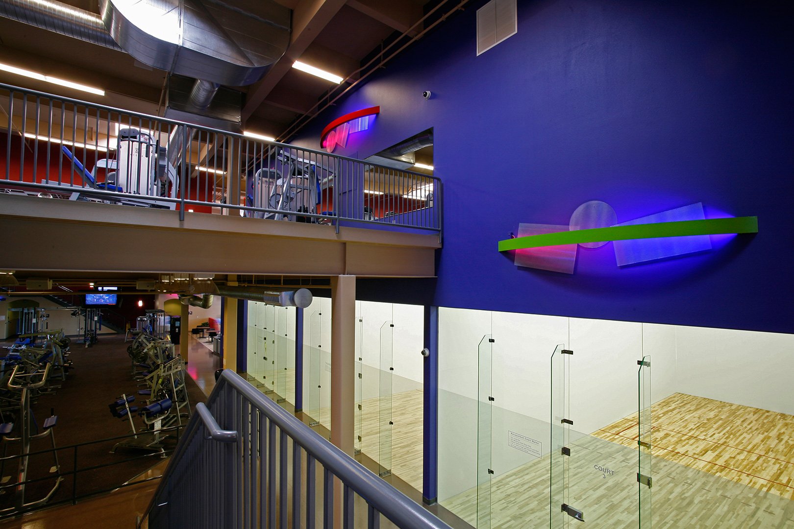 A racquetball court from the mezzanine through its glass back wall, its pale timber floor lit from above.