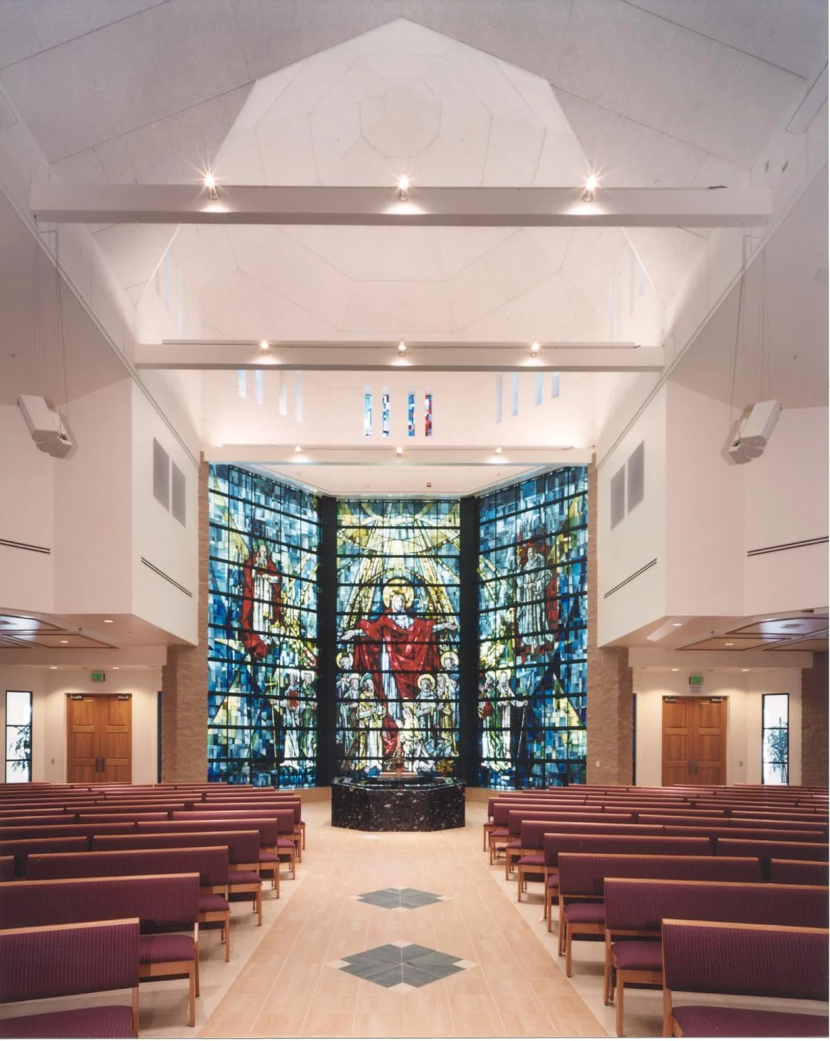 Down the centre aisle to the stained glass wall, a robed figure above a crowd of saints, with the stone font on the axis.