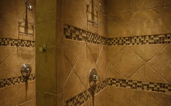 Two shower stalls divided by a tiled pier, travertine coloured tiles with a mosaic band at shoulder height.