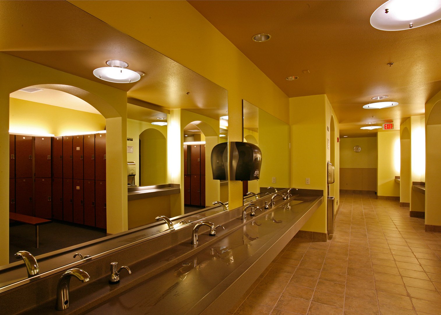 The washroom, a run of basins along a stainless counter under a long mirror reflecting the lockers.