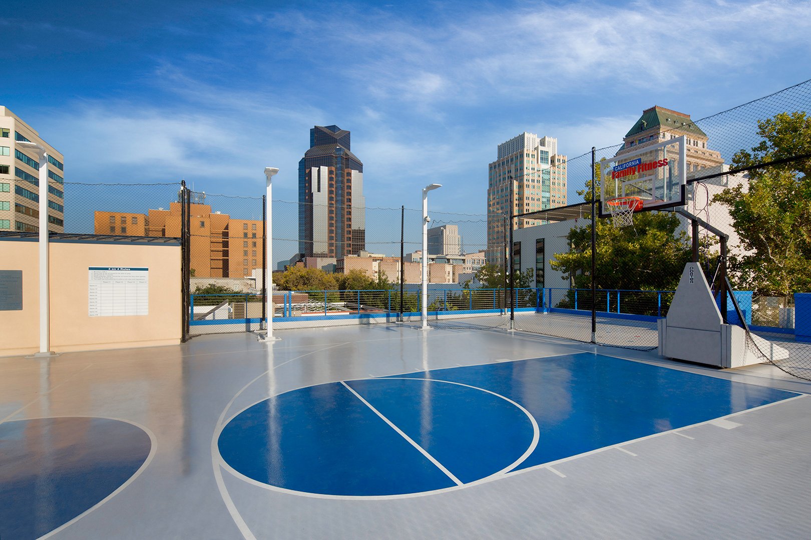 The rooftop court by day, a blue painted key on a pale deck inside chain link fencing, downtown towers beyond.