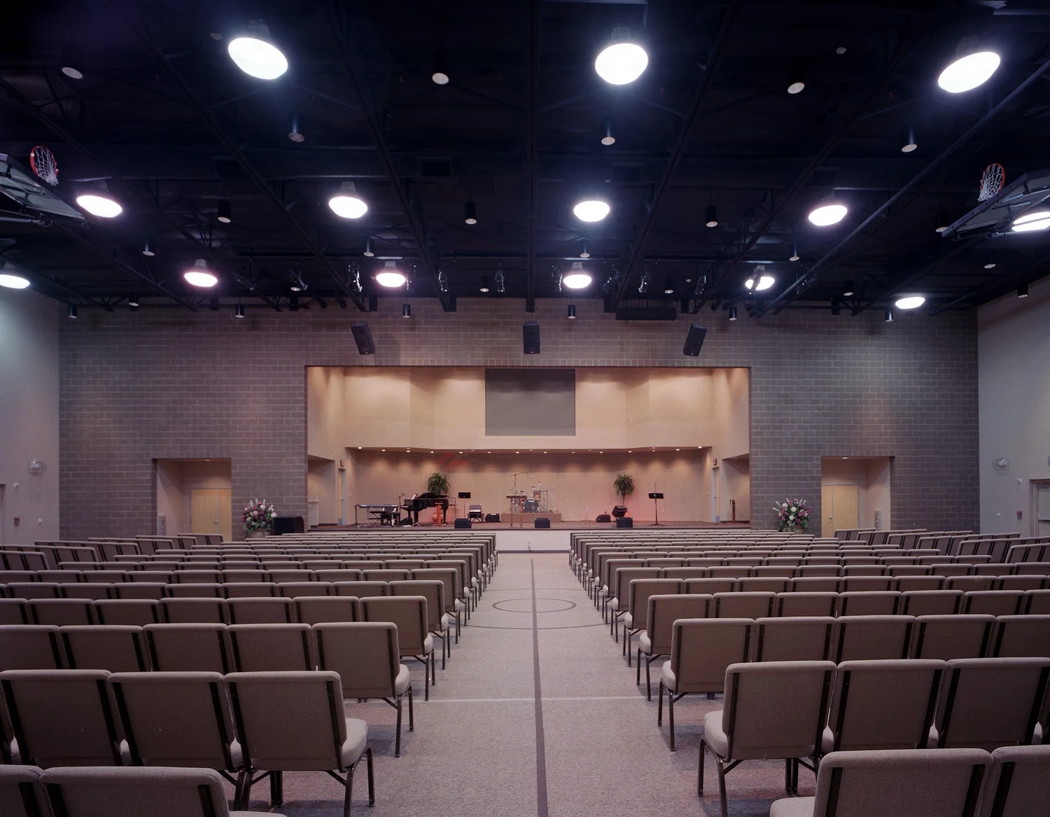 The multi-use hall set for worship, rows of chairs facing a low stage under exposed steel and folded basketball hoops.