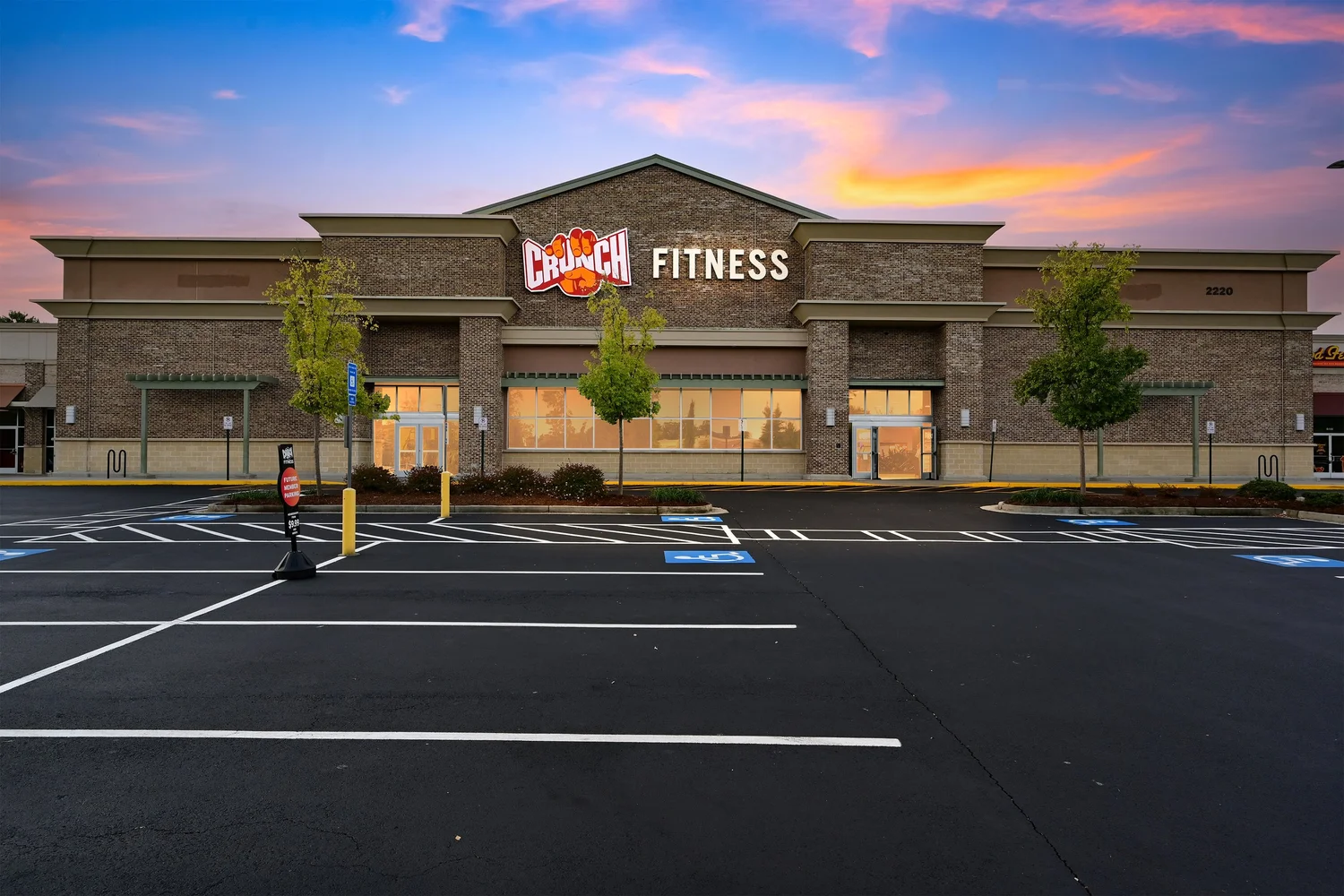 The club head on at sunset, a lit Crunch Fitness sign on the central gable above a brick frontage.