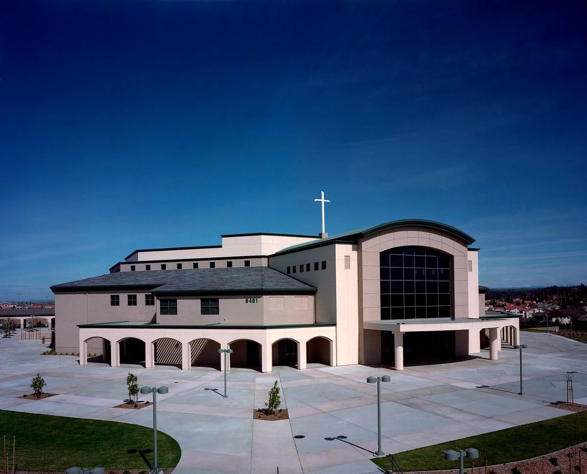 The same front closer in across the plaza, the arched arcade and the tall entrance window under a white cross.