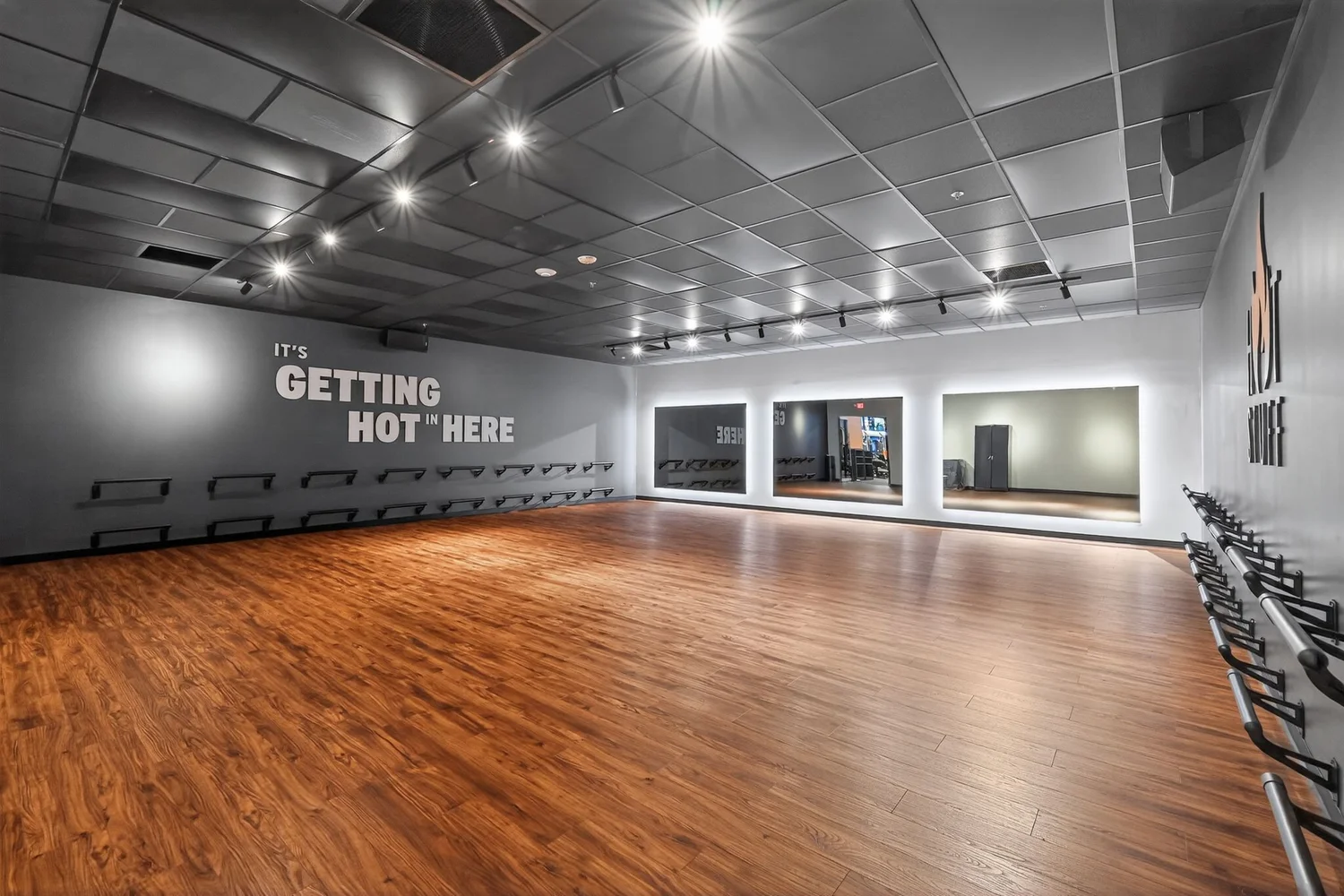An empty studio with a wood floor and mirrored end wall, It's Getting Hot In Here on the grey wall.