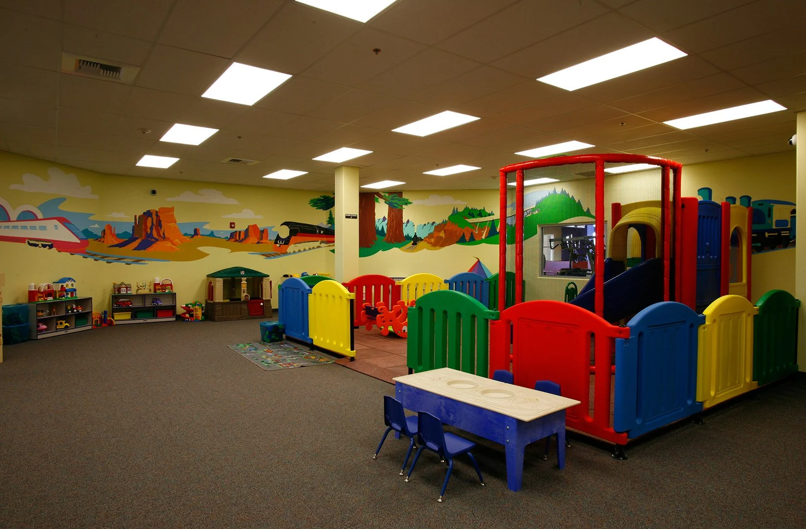 The child care room, primary coloured soft play behind a low gated frame, a painted mural along the wall.