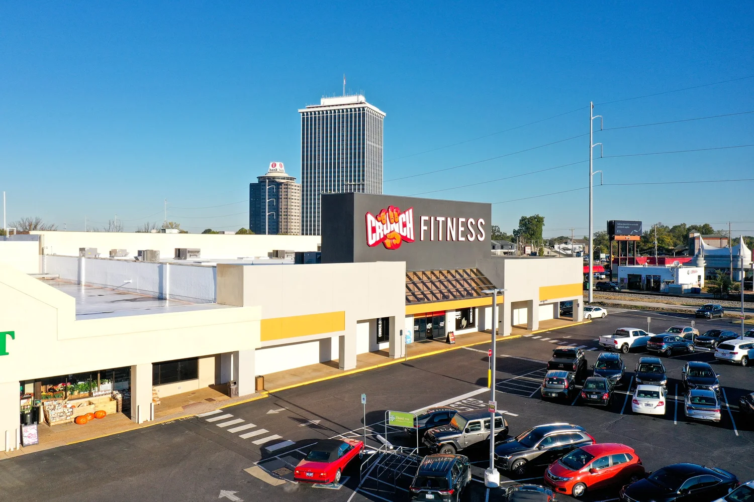 A cream strip centre with a dark sign block over its entrance, downtown office towers rising behind it.