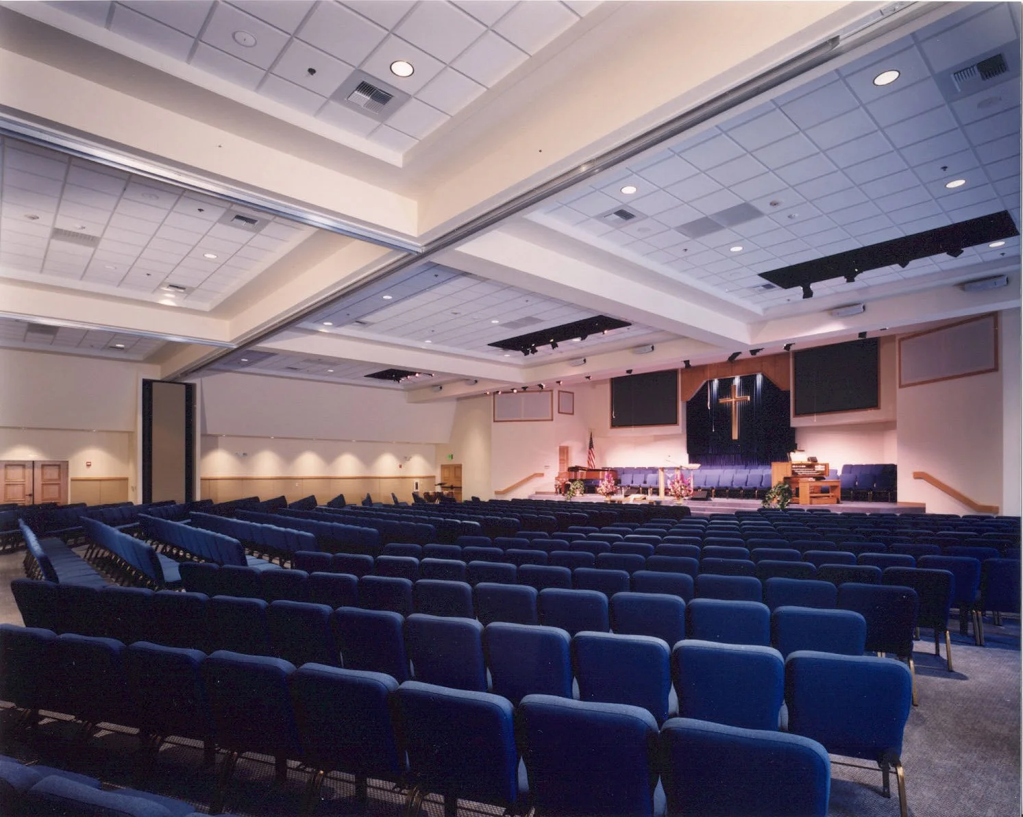 The sanctuary from the back, rows of blue chairs running down to a raised platform under a wide stepped ceiling.