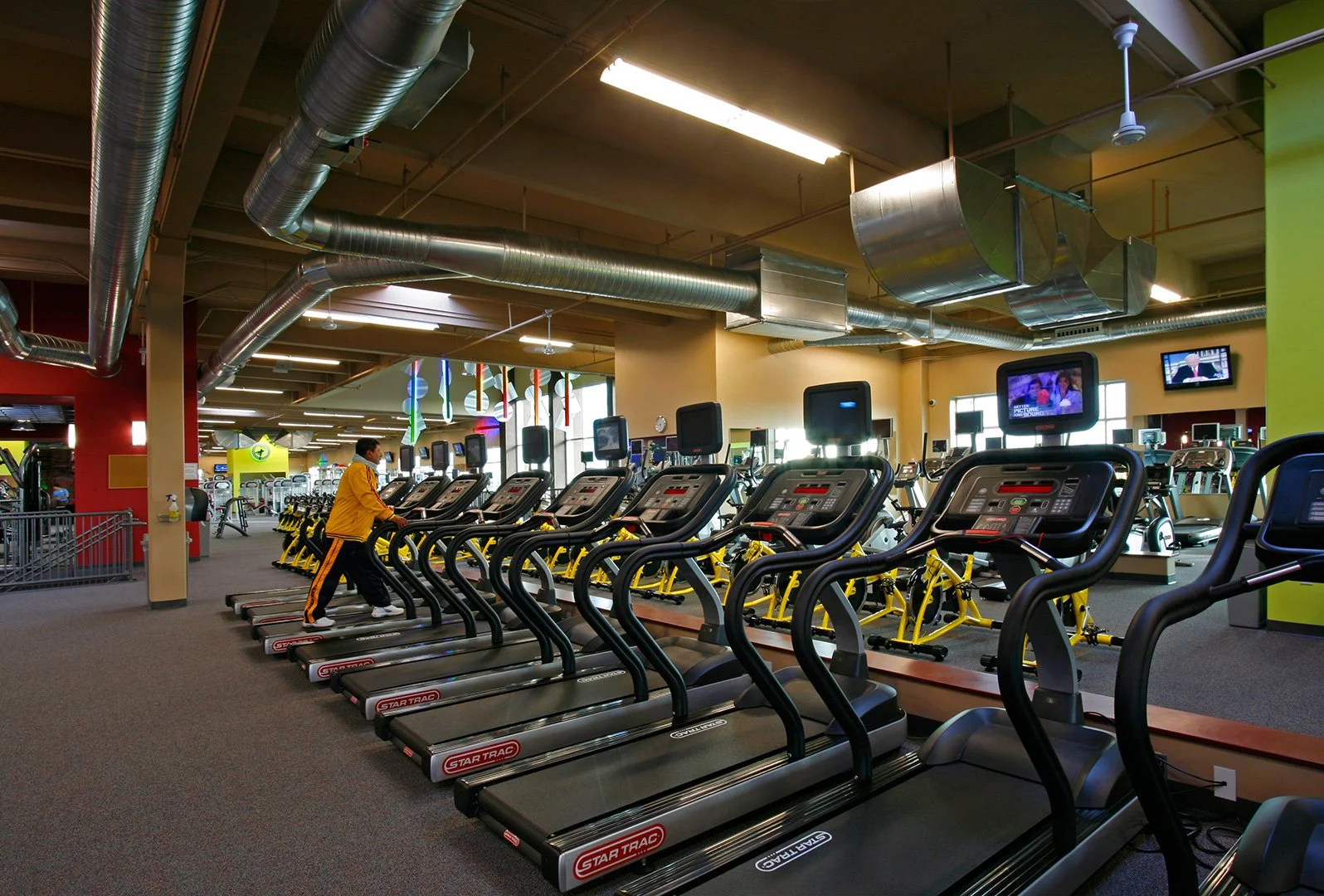Treadmills in a long row under an open timber roof and exposed ductwork, screens on brackets above.