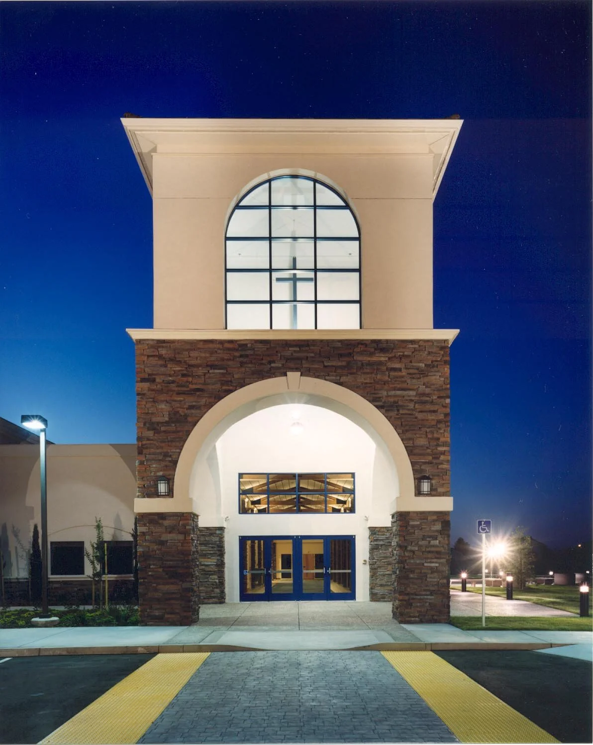 The entry tower head on after dark, a cross inside its lit arched window above a wide stone arch framing the glazed doors.