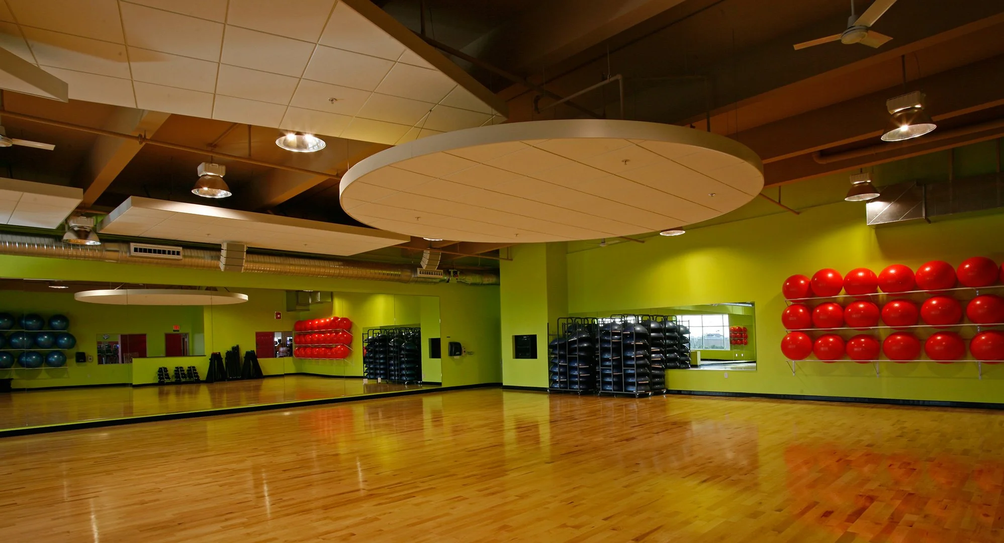 A group exercise studio, empty, a sprung timber floor running to a mirrored wall under a round acoustic disc.