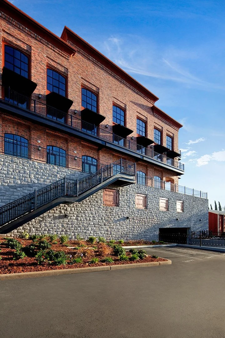 The downhill side, two brick storeys on a tall wall of pale blocks with an open steel stair descending across it.