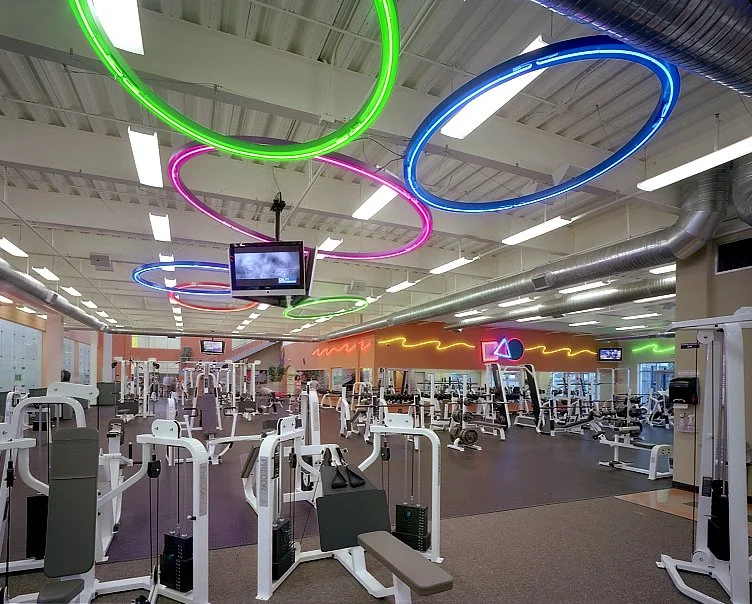 The weight floor from above, rows of machines and benches under green and pink neon rings.