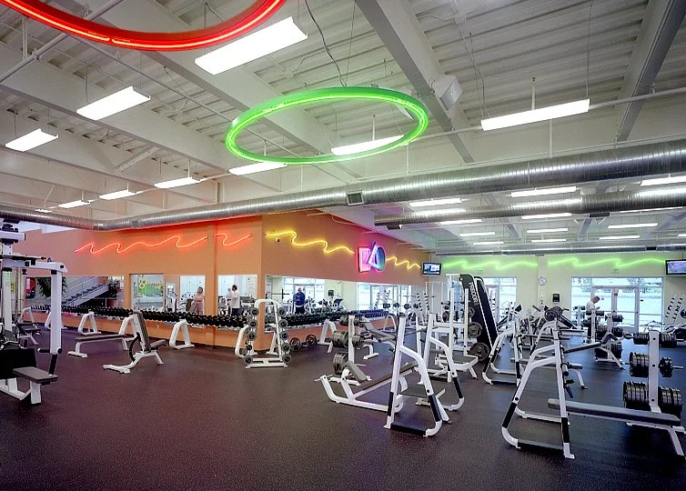 The cardio floor, ranks of treadmills and cross trainers facing a long orange wall under neon rings.