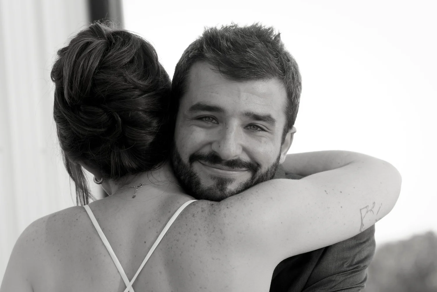 A black-and-white portrait of a couple embracing, the groom smiling