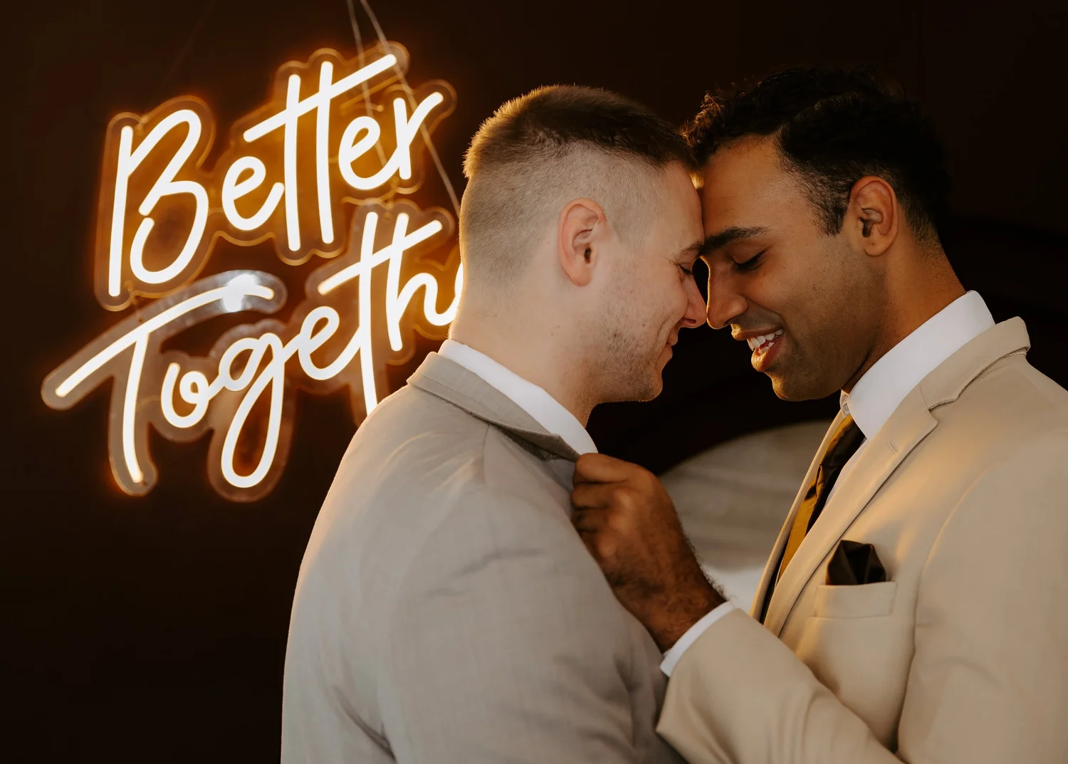 Two grooms in neutral suits sharing a close moment beneath a Better Together neon sign