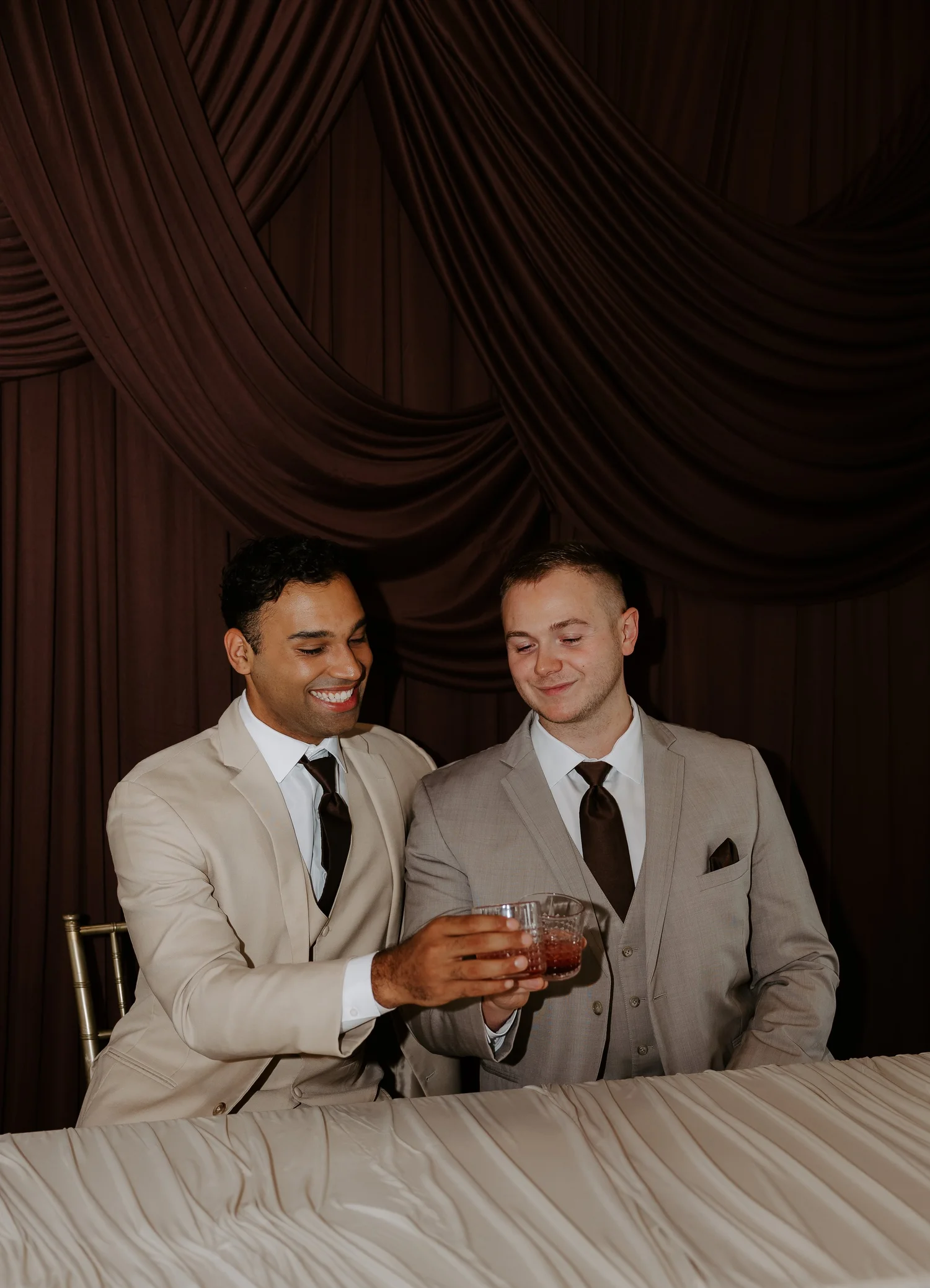 Two grooms in neutral suits seated together against chocolate draping