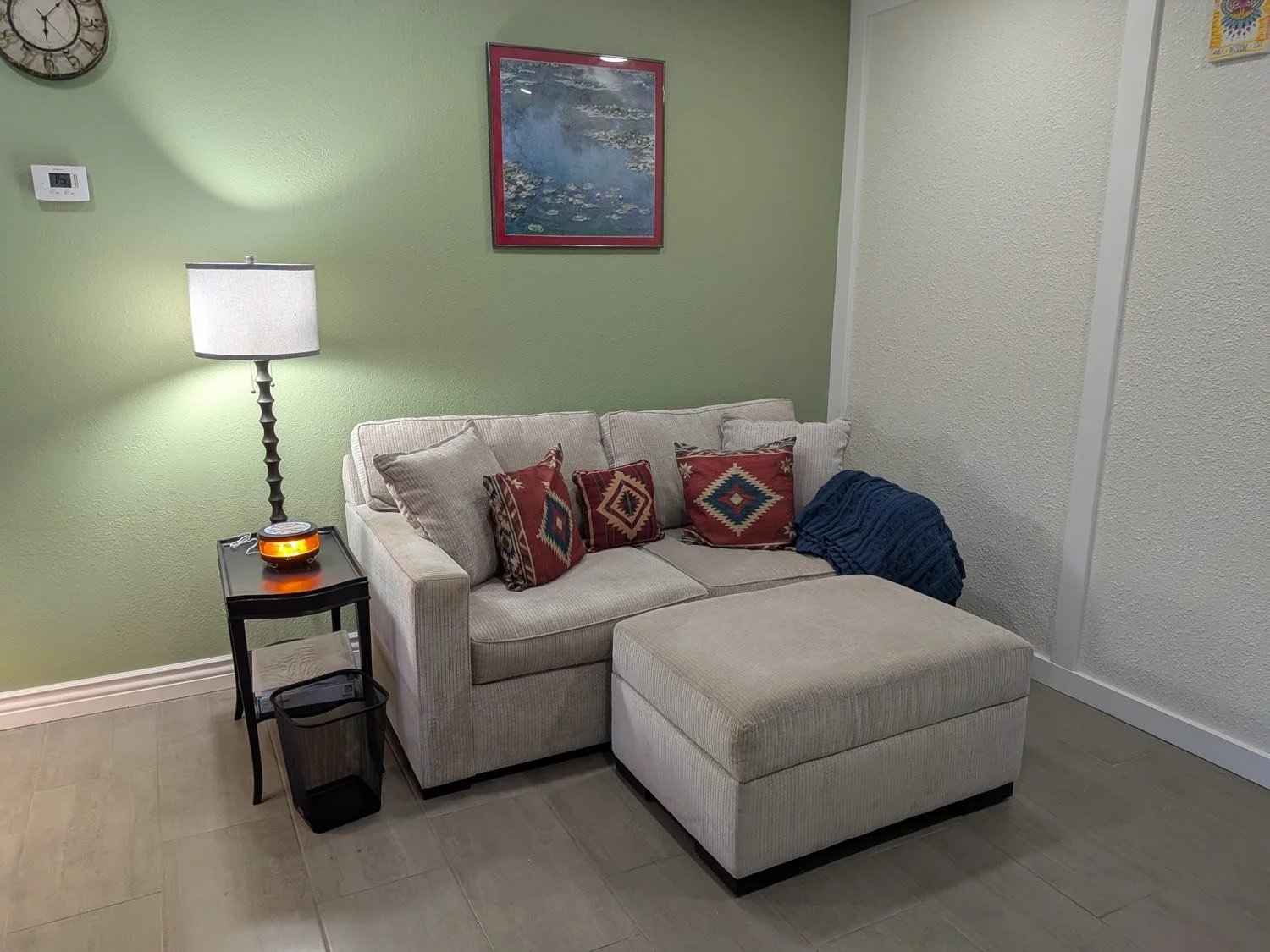 Inside Amy's Wickenburg therapy office — a soft sage-green room with a comfortable sofa, warm lamplight, Southwestern pillows, and a water-lily print on the wall