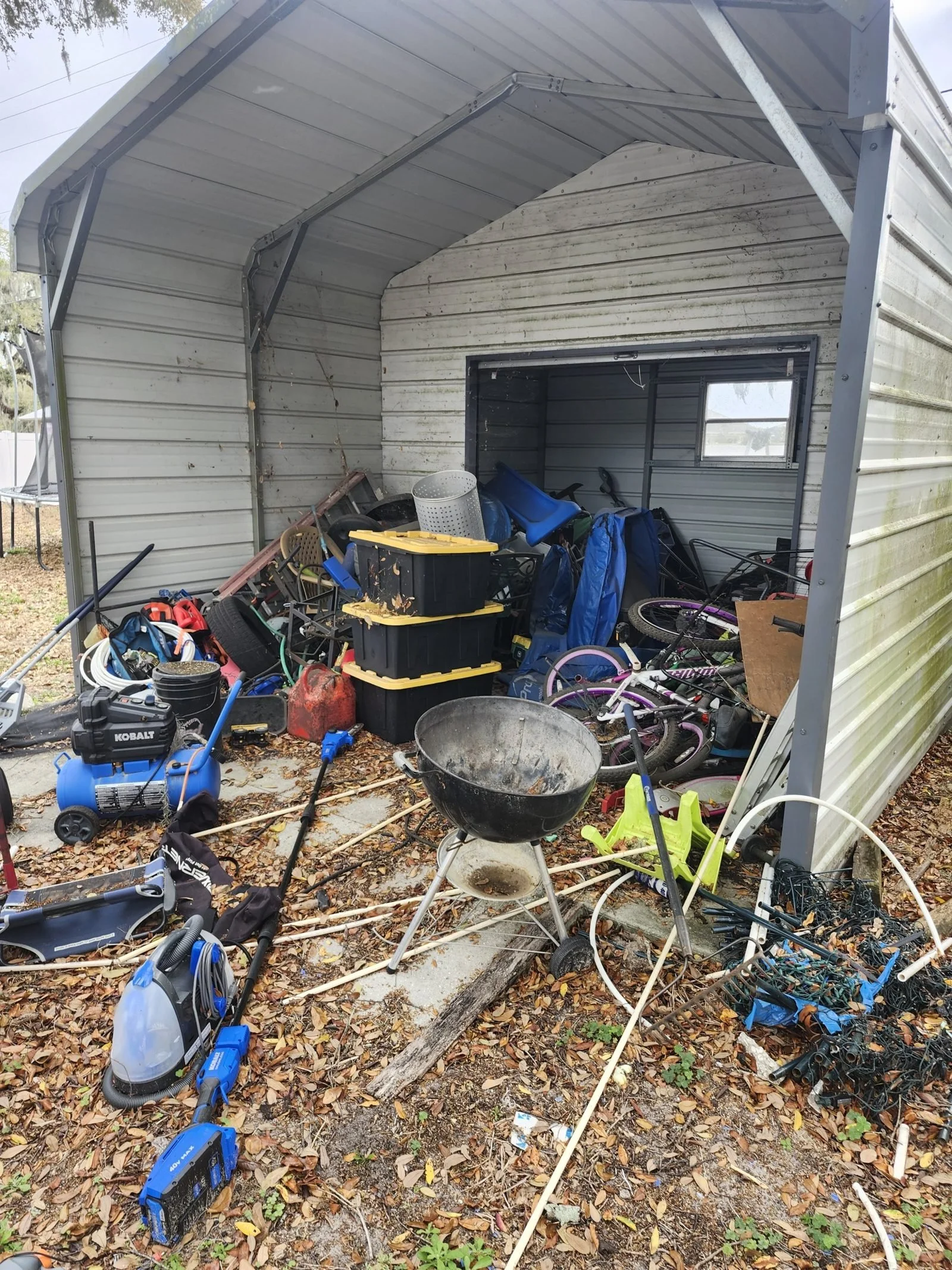 Metal carport stacked with bikes, bins and junk before the cleanout.
