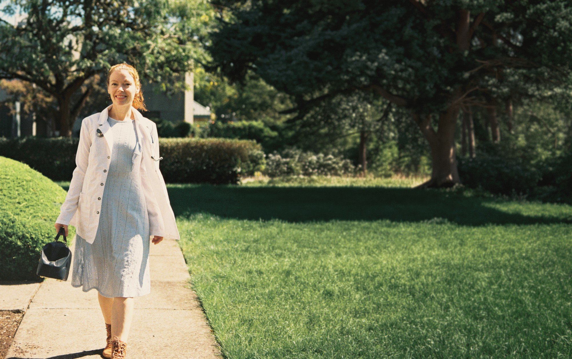 Dr. Abigail Stephens smiling in a white coat and carrying a doctor's bag, walking along a sunny garden path in Springfield, Oregon.