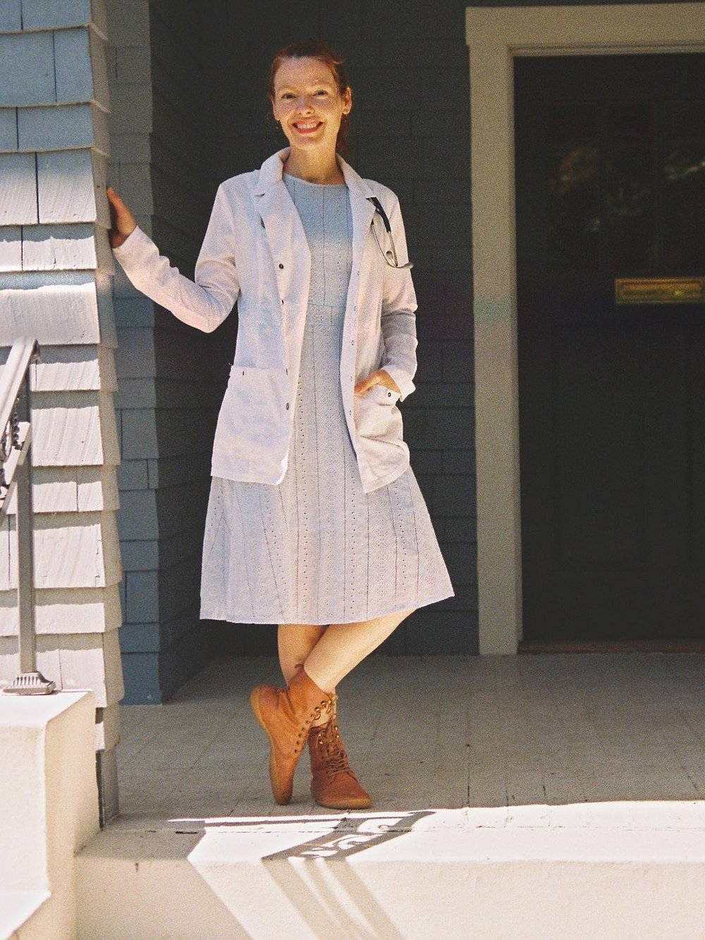Dr. Abigail Stephens in a white coat, smiling and leaning on the porch of the Stewart House in Springfield, Oregon, beside the navy front door.